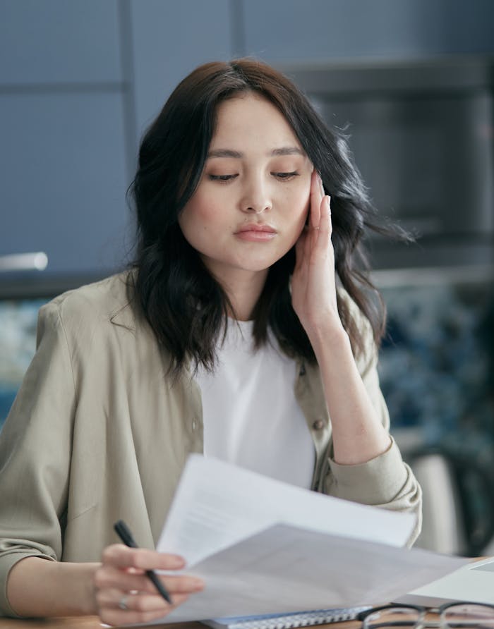 A thoughtful young woman holds papers and a pen, deeply engrossed in reading indoors.