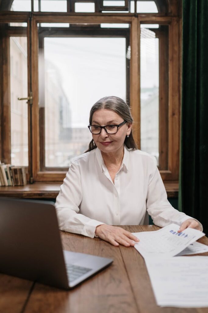 Elderly woman analyzing financial documents using a laptop indoors.