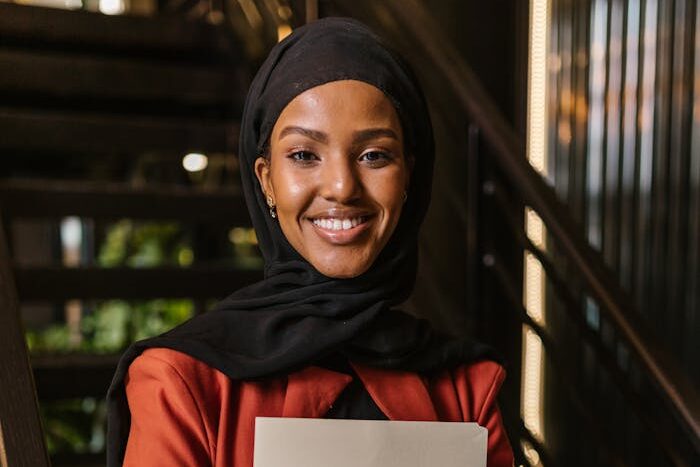 Professional woman in hijab holding folder, smiling in modern office setting.